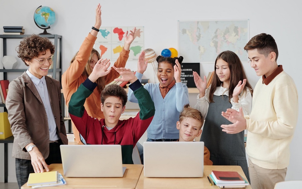 A group of children and teacher at desk excited, smiling, raising their hands, and clapping.
