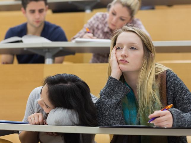 A person sleeping on a desk and bored looking student in tiered lecture theatre.
