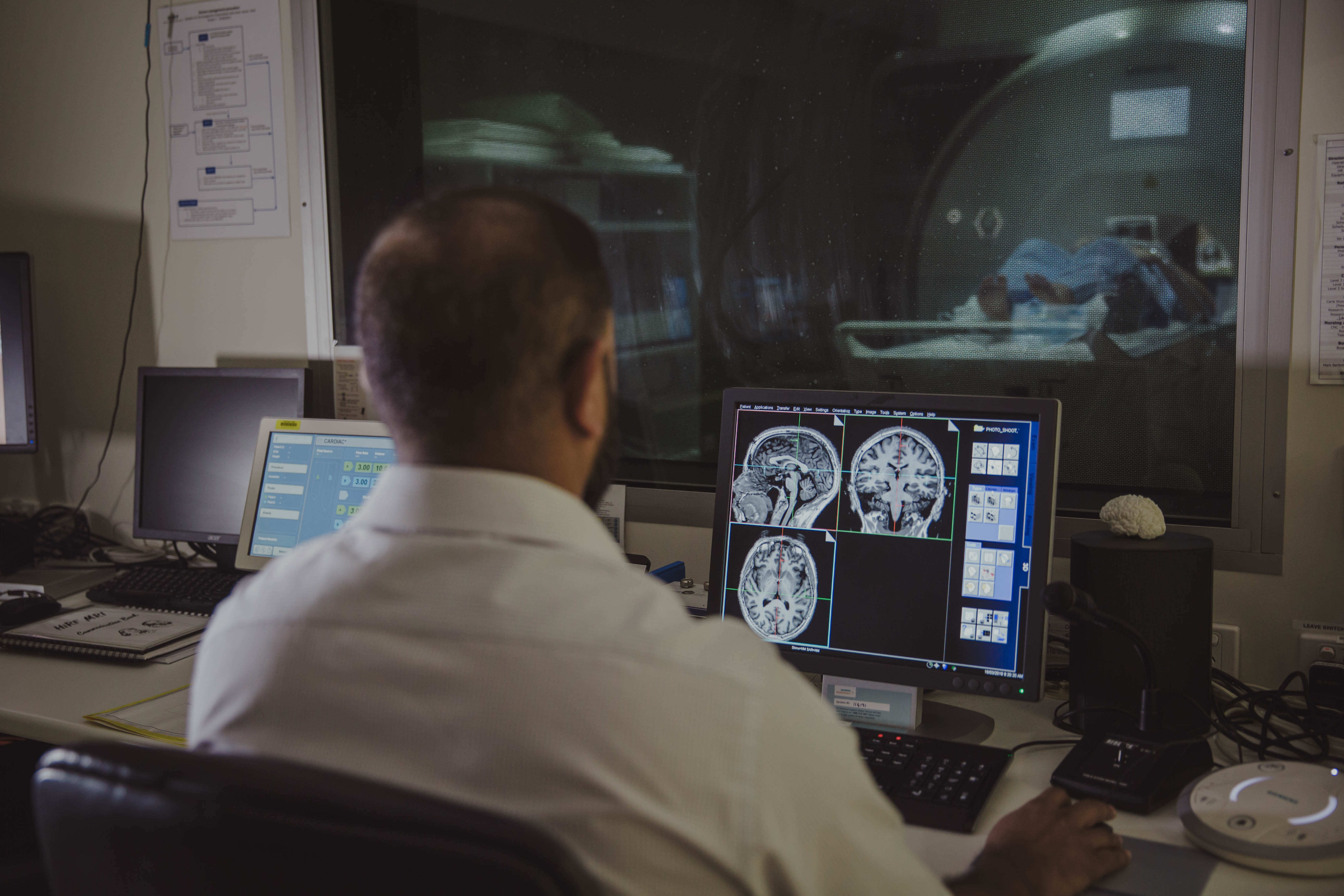 A person looking at computer screens displaying MRI brain scans. On the other side of the glass is the person being scanned in the MRI machine.