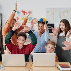 A group of children and teacher at desk excited, smiling, raising their hands, and clapping.