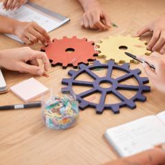 A group of people collaborating on a table with notepads, pens, and gears.