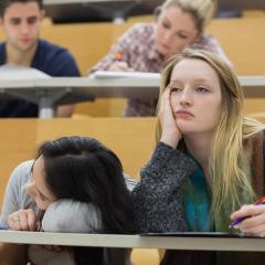 A person sleeping on a desk and bored looking student in tiered lecture theatre.